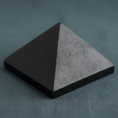 Close-up view of a black shungite pyramid showing natural surface texture on a muted teal linen background with soft diffused lighting. — for Carpe Diem Dream