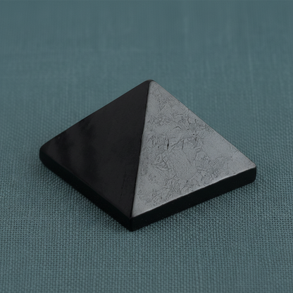 Black shungite pyramid displayed upright on a muted teal linen surface with soft diffused lighting and subtle shadow. — for Carpe Diem Dream