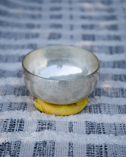 Silver Tibetan singing bowl with engraved mantra symbols resting on a yellow embroidered cushion on a textured woven cloth surface. — for Carpe Diem Dream