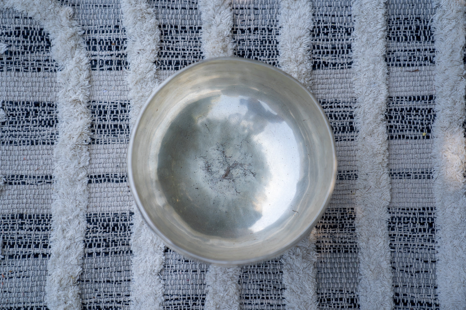 Close-up top view of a silver Tibetan singing bowl showing the hammered interior surface resting on a textured woven cloth background. — for Carpe Diem Dream