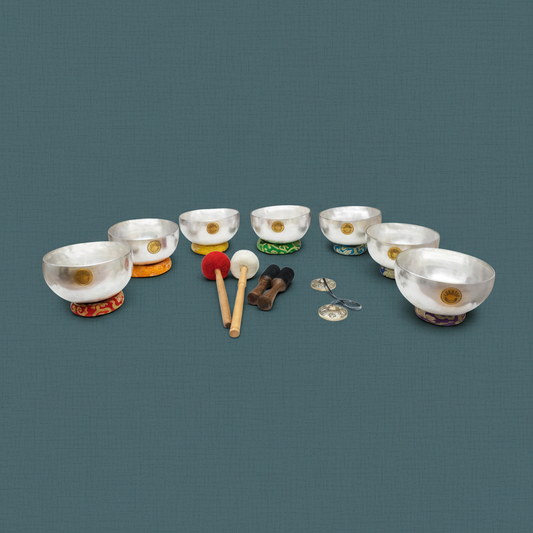 Seven silver Tibetan singing bowls arranged in a semicircle with embroidered chakra cushions, wooden mallets, and tingsha cymbals on a muted teal textured background. — for Carpe Diem Dream