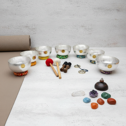 Seven silver Tibetan singing bowls arranged in a semicircle with embroidered chakra cushions, wooden mallets, tingsha cymbals, and colorful chakra stones with a tan yoga mat on a clean white background. — for Carpe Diem Dream 
