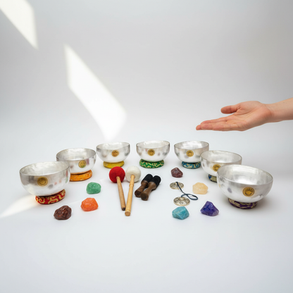 Seven silver Tibetan singing bowls arranged in a semicircle with embroidered chakra cushions, wooden mallets, tingsha cymbals, and colorful chakra stones with a hand shown above for scale on a clean white background. — for Carpe Diem Dream