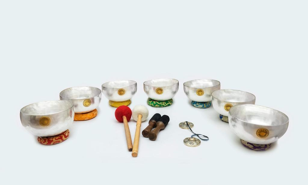 Seven silver Tibetan singing bowls with embroidered chakra cushions arranged with mallets, rotating sticks, and tingshaw cymbals on a white background. — for Carpe Diem Dream
