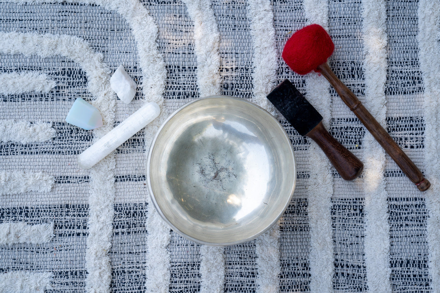 Top view of a silver Tibetan singing bowl with wooden mallets and small ritual stones arranged on a textured woven cloth surface. — for Carpe Diem Dream
