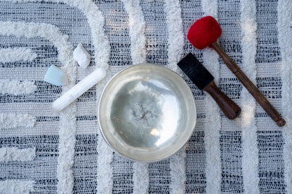 Top view of a silver Tibetan singing bowl with wooden mallets and small ritual stones arranged on a textured woven cloth surface. — for Carpe Diem Dream