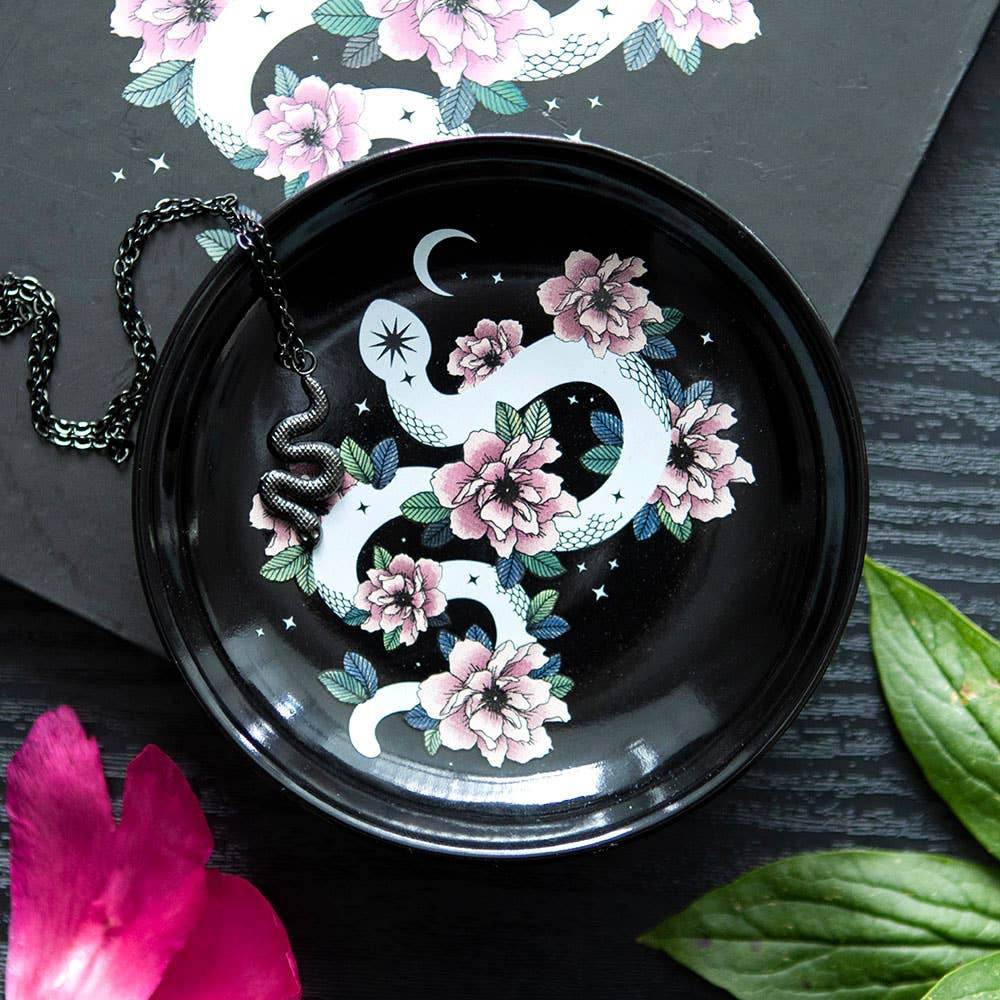 Styled photo of Snake & Flower Celestial Trinket Dish on a dark wooden surface with jewelry and leaves, showing white serpent and pink flowers under soft lighting in Carpe Diem Dream’s mystical, gothic-inspired décor aesthetic.