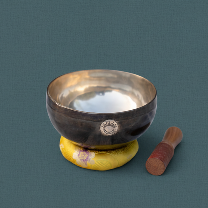 A Solar Plexus Chakra singing bowl with wooden striker resting on a yellow cushion against a muted teal linen background. — for Carpe Diem Dream