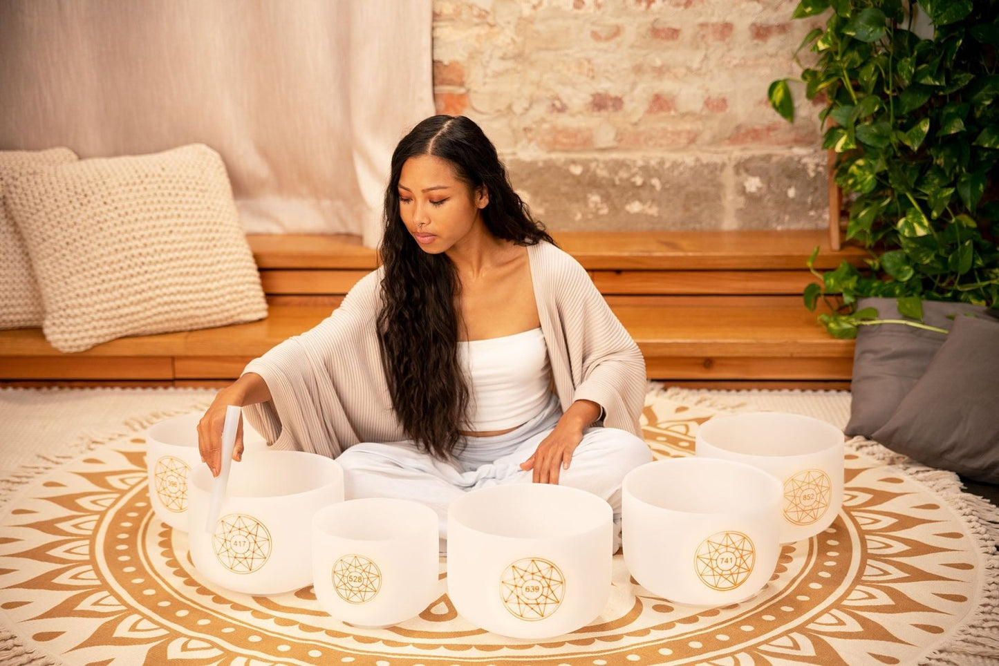 Woman seated on a patterned rug arranging six white frosted crystal singing bowls with gold frequency symbols in a circular formation — for Carpe Diem Dream