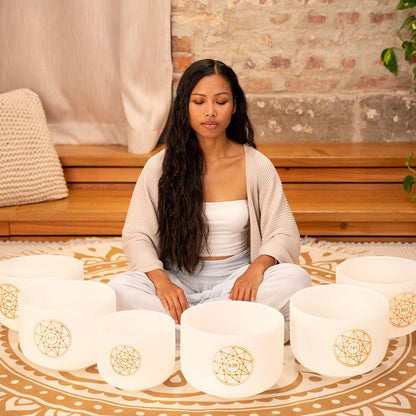 Woman seated in meditation surrounded by six white frosted crystal singing bowls with gold frequency symbols arranged in a circle on a patterned rug — for Carpe Diem Dream