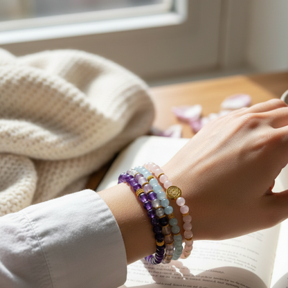 A woman’s wrist wearing stacked gemstone bracelets rests on an open book beside a soft knit blanket in warm natural light — for Carpe Diem Dream