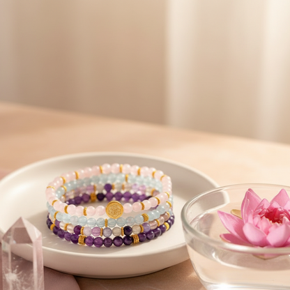 A stacked set of gemstone bracelets rests inside a shallow ceramic dish beside a glass bowl with a floating pink lotus on a blush surface in soft ambient light. — for Carpe Diem Dream