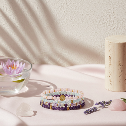 A stacked set of pastel gemstone bracelets displayed on a blush satin surface beside a glass bowl with a floating lotus and soft botanical shadows. — for Carpe Diem Dream