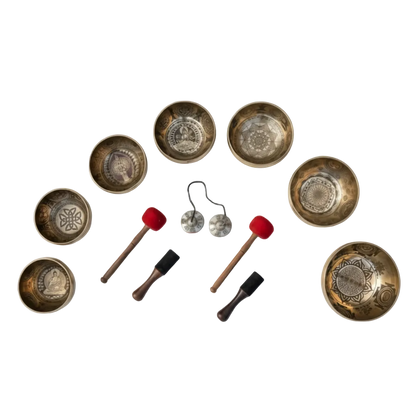 Seven engraved Tibetan singing bowls with carved chakra symbols arranged with wooden mallets and tingsha meditation bells on a clean background. — for Carpe Diem Dream