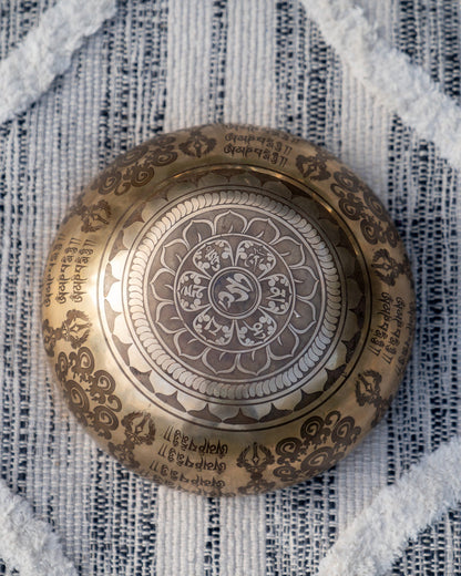 Bottom view of carved Tibetan singing bowl showing Om symbol within an intricate mandala engraving on textured blue and white linen fabric. — for Carpe Diem Dream