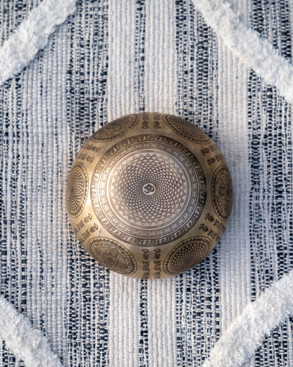 Bottom view of carved Tibetan singing bowl showing Om symbol within a radiating mandala engraving on textured blue and white linen fabric. — for Carpe Diem Dream