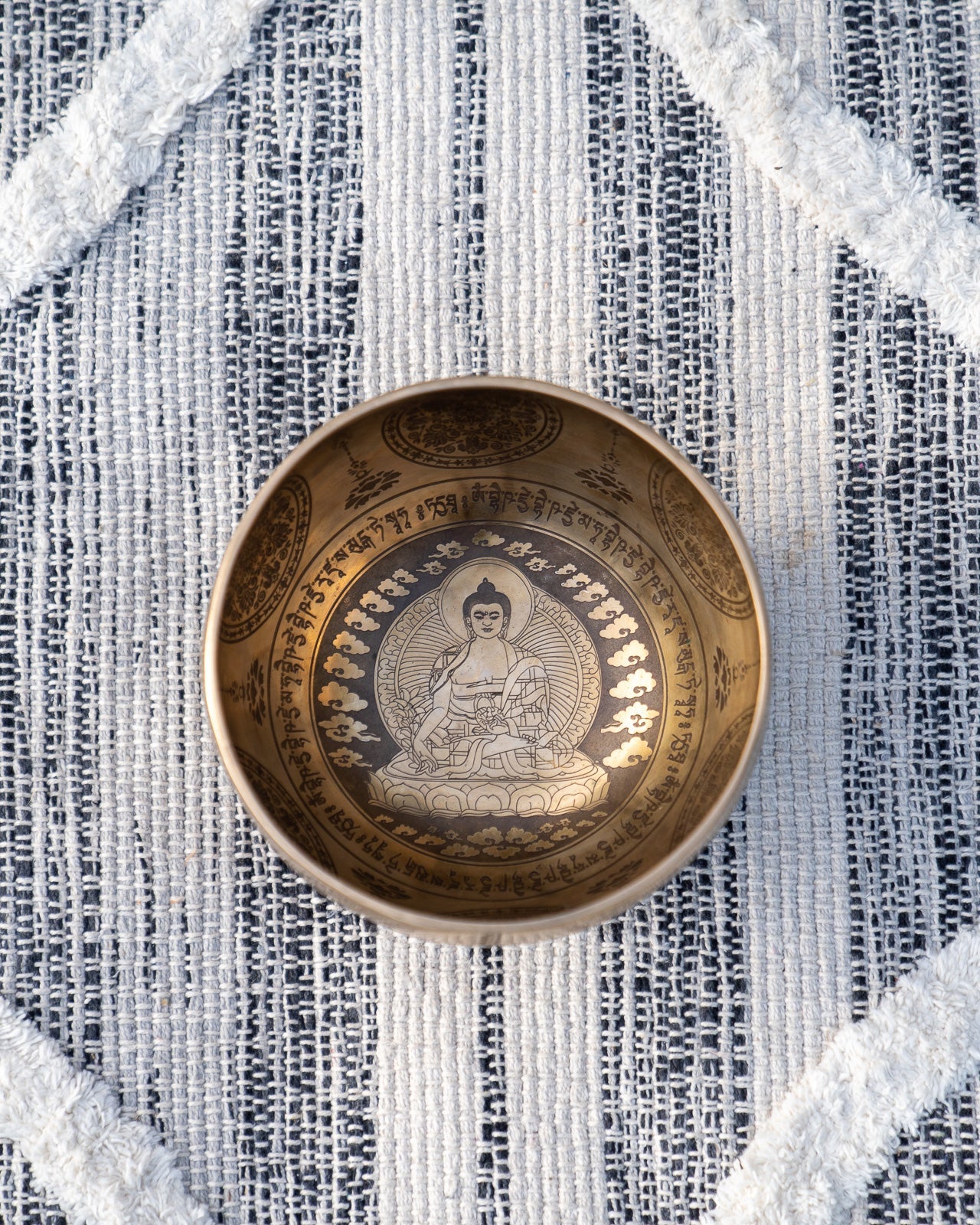 Top view of carved Tibetan singing bowl showing Buddha meditating within a halo mandala surrounded by mantra script on textured blue and white linen fabric. — for Carpe Diem Dream