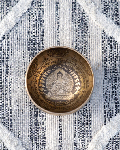 Top view of carved Tibetan singing bowl showing Buddha meditating within a halo mandala surrounded by mantra script on textured blue and white linen fabric. — for Carpe Diem Dream