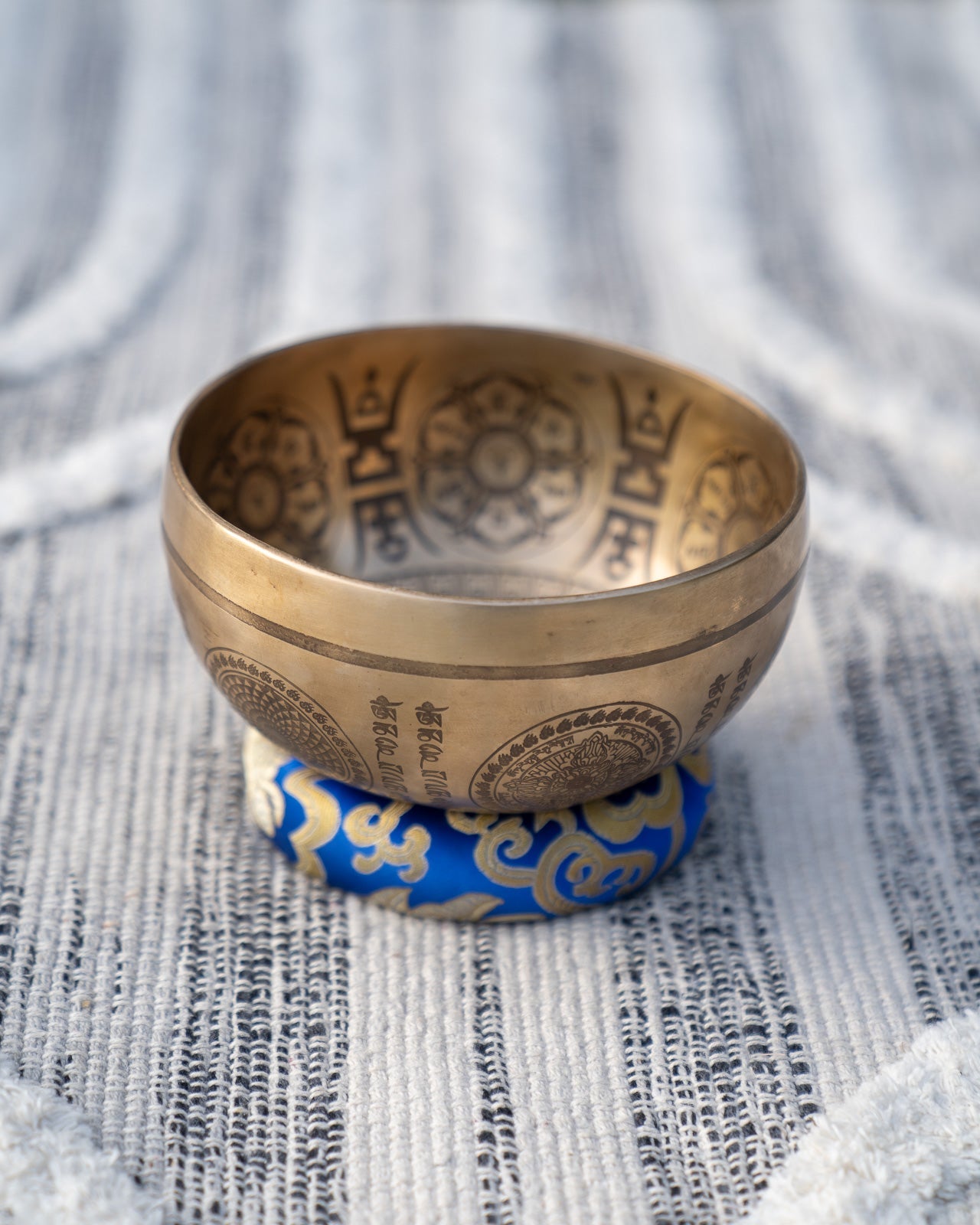 Carved Tibetan singing bowl with mandala engravings resting on a purple crown chakra cushion on textured blue and white linen outdoors. — for Carpe Diem Dream