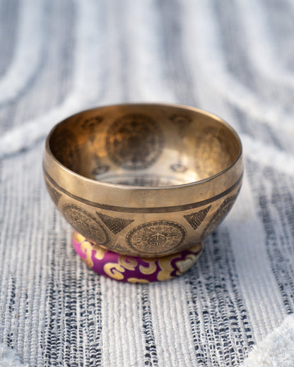 Carved Tibetan singing bowl with mandala engravings resting on a purple crown chakra cushion on textured blue and white linen outdoors. — for Carpe Diem Dream