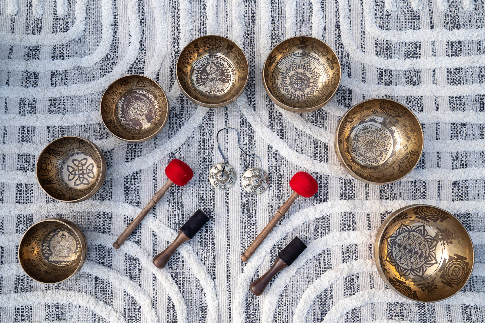 Top view of seven carved Tibetan singing bowls with embroidered chakra cushions, mallets, rotating sticks, and tingsha cymbals arranged on textured blue and white linen. — for Carpe Diem Dream