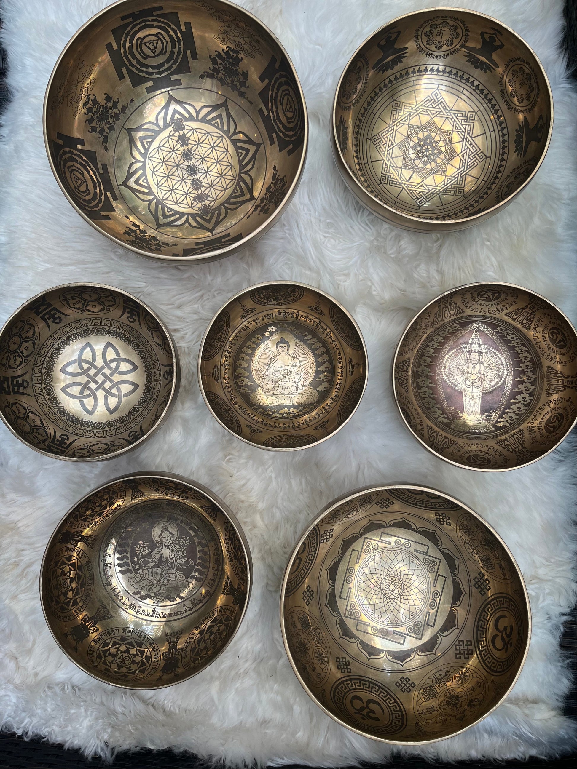 Top view of seven carved Tibetan singing bowls with embroidered chakra cushions, mallets, rotating sticks, and tingsha cymbals arranged on a white fir surface. — for Carpe Diem Dream