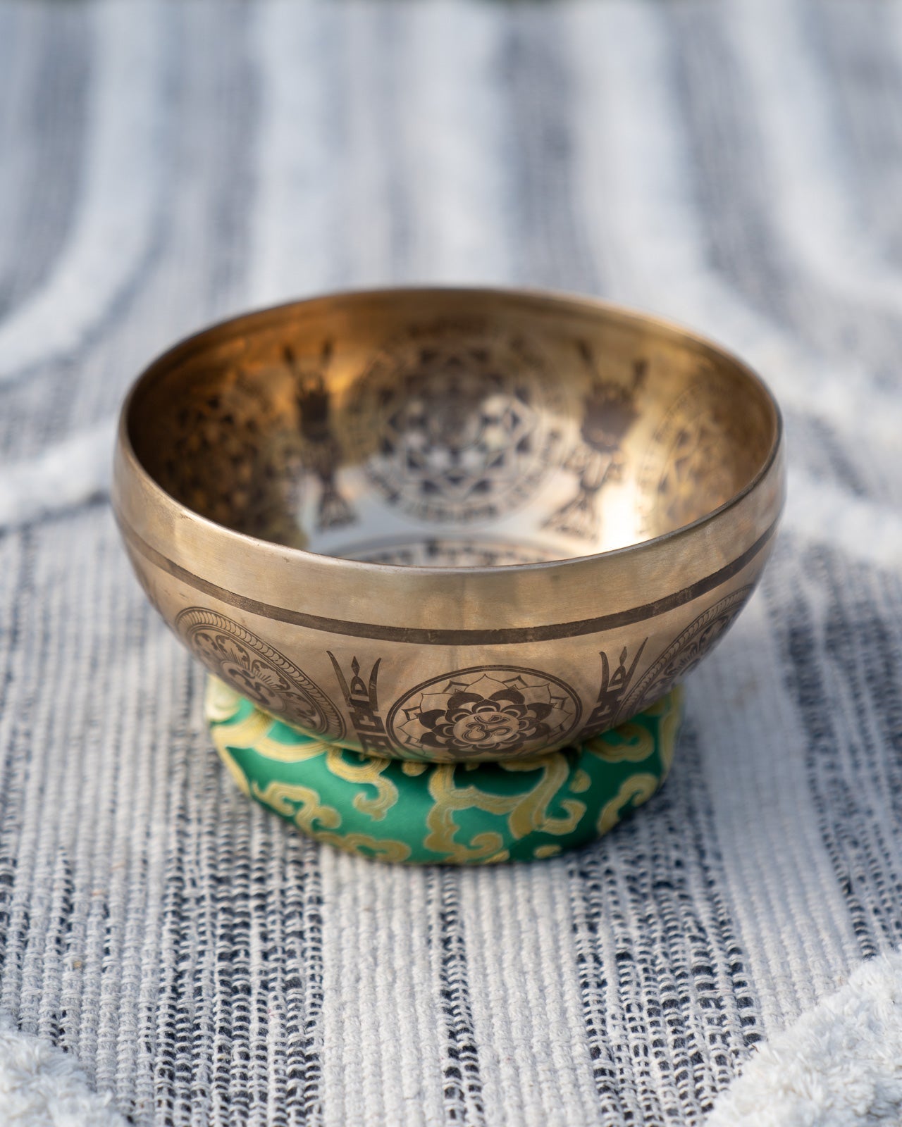 Carved Tibetan singing bowl with mandala engravings resting on a green heart chakra cushion on textured blue and white linen outdoors. — for Carpe Diem Dream