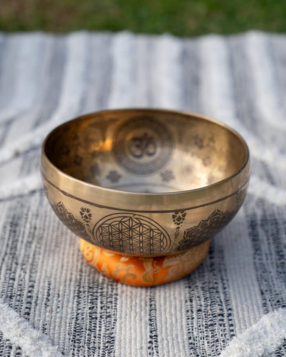 Carved Tibetan singing bowl with mandala engravings resting on an orange sacral chakra cushion on textured blue and white linen outdoors. — for Carpe Diem Dream