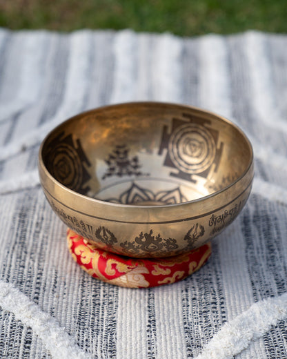 Single carved Tibetan singing bowl with Sanskrit and mandala engravings resting on a red embroidered cushion on textured blue and white linen outdoors. — for Carpe Diem Dream