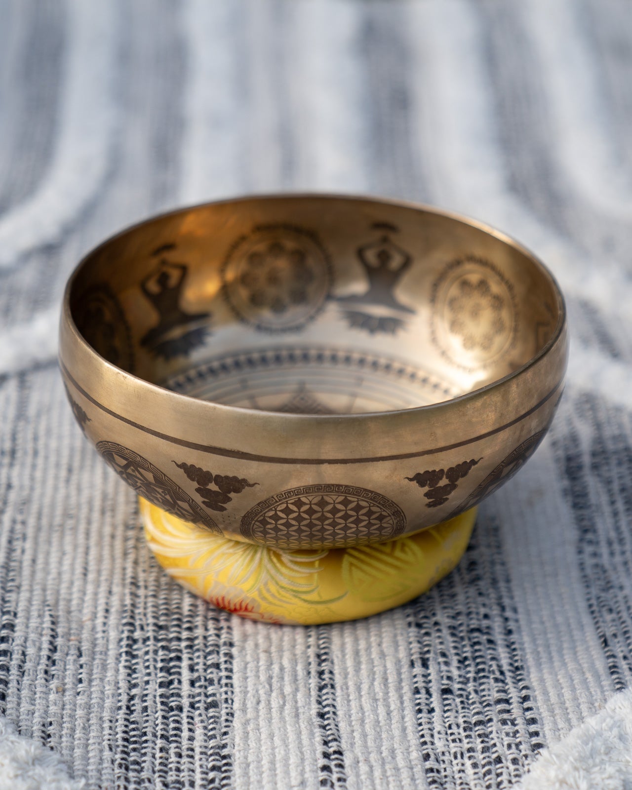 Carved Tibetan singing bowl with mandala engravings resting on a yellow solar plexus chakra cushion on textured blue and white linen outdoors. — for Carpe Diem Dream