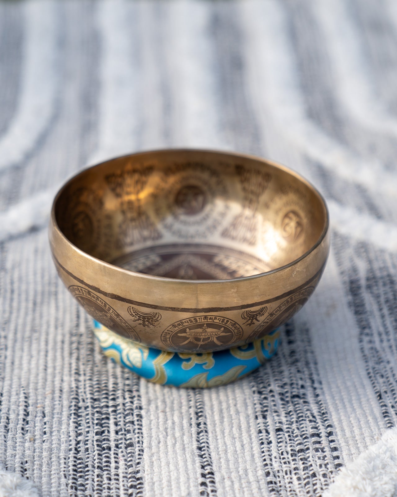 Carved Tibetan singing bowl with mandala engravings resting on a blue throat chakra cushion on textured blue and white linen outdoors. — for Carpe Diem Dream
