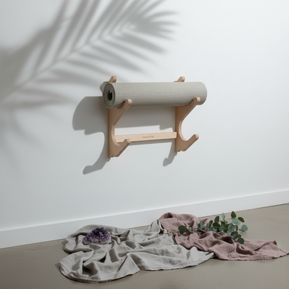 Natural wood StackRack yoga prop wall organizer holding a gray yoga mat on a white wall with palm leaf shadows, gray and mauve fabric, eucalyptus, and an amethyst crystal below. — for Carpe Diem Dream