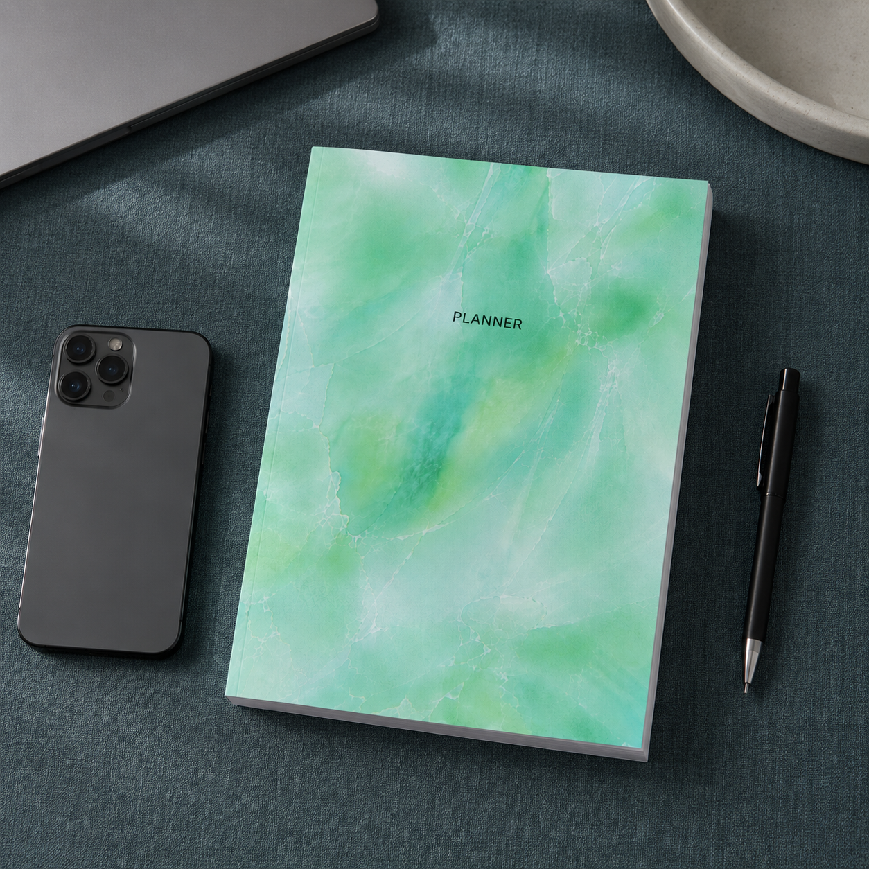Start Anytime Undated 52-Week Planner paperback by Big Fish Planners shown from an overhead perspective on a #374F59 textured linen surface beside a smartphone and pen for realistic size comparison, featuring the original mint watercolor cover design, accurate paperback proportions, visible page block, and cool editorial studio lighting — for Carpe Diem Dream