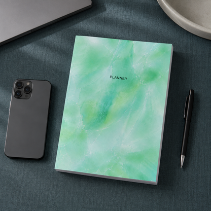 Start Anytime Undated 52-Week Planner paperback by Big Fish Planners shown from an overhead perspective on a #374F59 textured linen surface beside a smartphone and pen for realistic size comparison, featuring the original mint watercolor cover design, accurate paperback proportions, visible page block, and cool editorial studio lighting — for Carpe Diem Dream