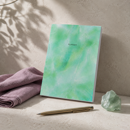 Start Anytime Undated 52-Week Planner paperback by Big Fish Planners displayed in the Carpe Diem Dream signature studio botanical setting with cool limestone textures, a soft botanical shadow, folded mauve textile accent, polished green crystal, and elegant brass pen, photographed in diffused natural light while preserving the exact planner cover artwork and realistic softcover paperback construction.
