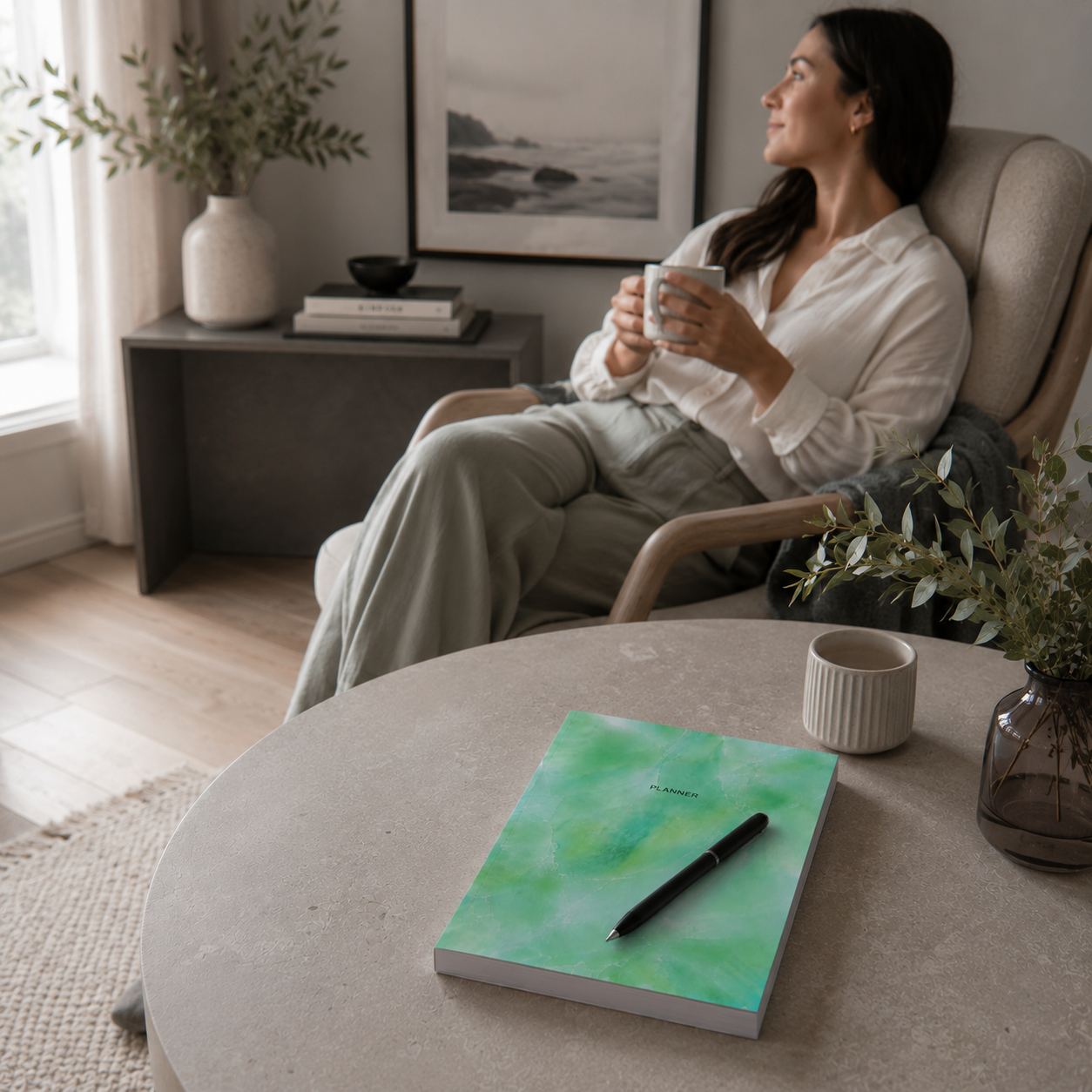 Start Anytime Undated 52-Week Planner paperback by Big Fish Planners resting closed with a pen placed across the cover on a cool limestone coffee table, while a relaxed woman enjoys a quiet moment with tea in an organized living space, illustrating clarity, peace of mind, reduced stress, and the feeling of completion after a productive planning session — for Carpe Diem Dream