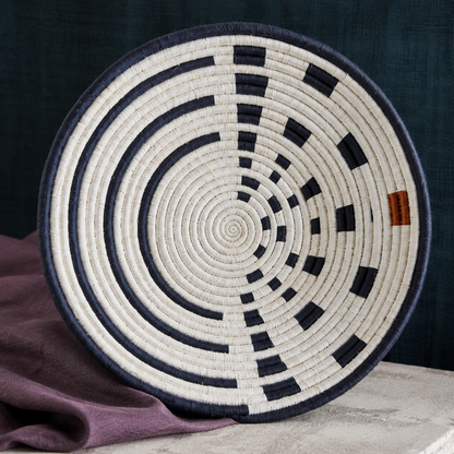 Close-up of large Sunrise Wall Basket handwoven natural grass and navy thread detail styled with mauve linen, dark teal backdrop, and soft stone surface. — for Carpe Diem Dream