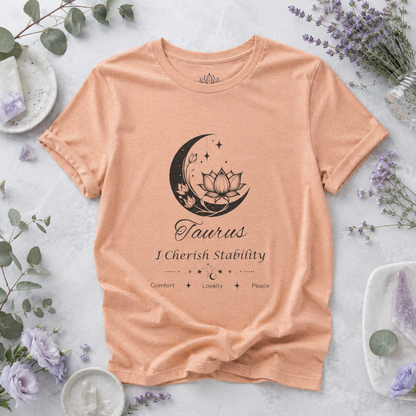 Taurus Moon I Cherish Stability zodiac T-shirt in Heather Peach with black moon-inspired lotus, floral, and celestial artwork and Comfort, Loyalty, Peace text, styled in a cool-toned studio botanical flat lay with eucalyptus, lavender flowers, crystals, and ceramic accents. — for Carpe Diem Dream