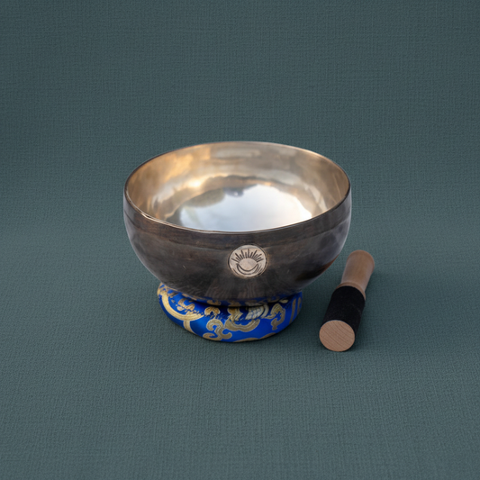 Hand-hammered Third Eye Chakra New Moon singing bowl tuned to A note resting on a blue cushion with wooden mallet on a muted teal linen background. — for Carpe Diem Dream