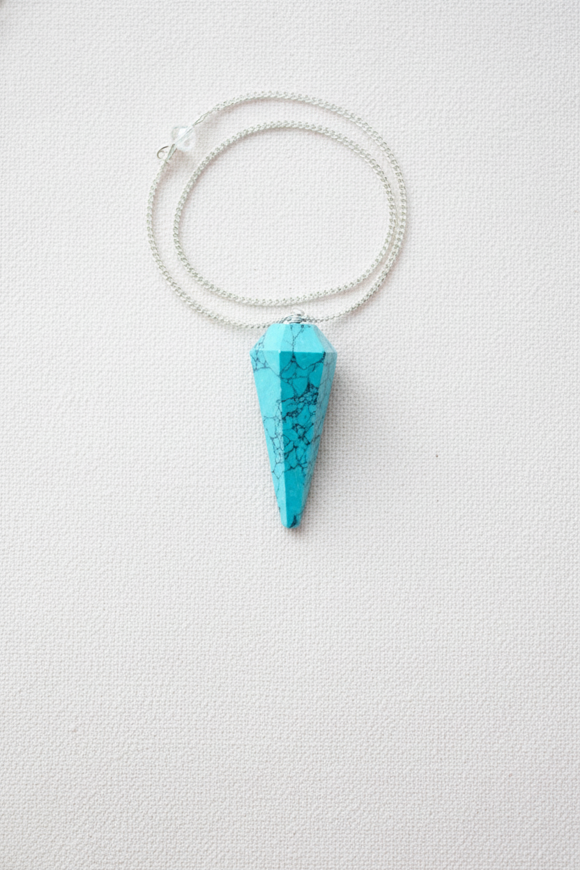 Turquoise crystal pendulum with silver chain displayed on a white textured surface, highlighting its vibrant blue tone and natural veining – for Carpe Diem Dream.