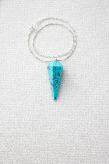 Turquoise crystal pendulum with silver chain displayed on a white textured surface, highlighting its vibrant blue tone and natural veining – for Carpe Diem Dream.