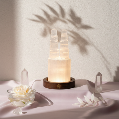 Two tower selenite crystal lamp glowing on a wooden base in a soft botanical studio setting with crystal points and white flowers. — for Carpe Diem Dream