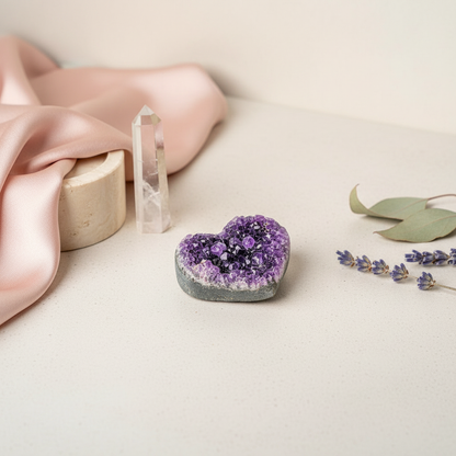 A heart-shaped Uruguayan amethyst cluster displayed on a light stone surface with soft pink fabric, clear quartz points, and lavender sprigs in a serene studio ritual setting. — for Carpe Diem Dream