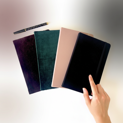 Four velvet hard cover journals in maroon, forest green, blush pink, and black fanned out beside a hand pointing to one journal to show size and scale. — for Carpe Diem Dream