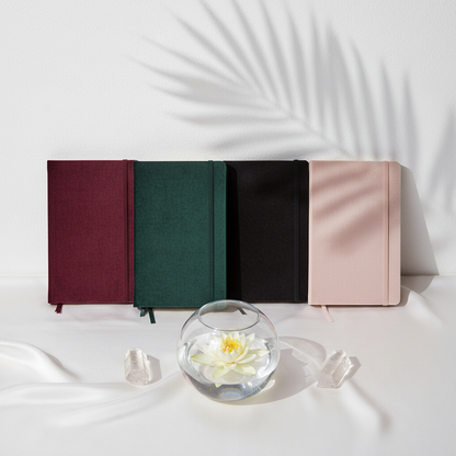 Four velvet hard cover journals in maroon, forest green, black, and blush pink arranged beside a lotus flower in a glass bowl and clear crystals in a soft botanical setting. — for Carpe Diem Dream
