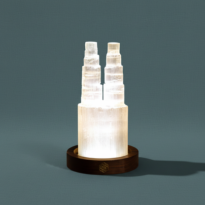 Two tower selenite crystal lamp glowing on a round wooden base against a muted teal background. — for Carpe Diem Dream