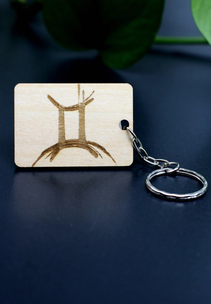 Wood Engraved Zodiac Keychain featuring Gemini astrology symbol, handcrafted from natural wood blue background with green plant for Carpe Diem Dream