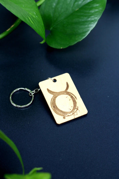 Wood Engraved Zodiac Keychain featuring Taurus astrology symbol, handcrafted from natural wood blue background with green plant for Carpe Diem Dream