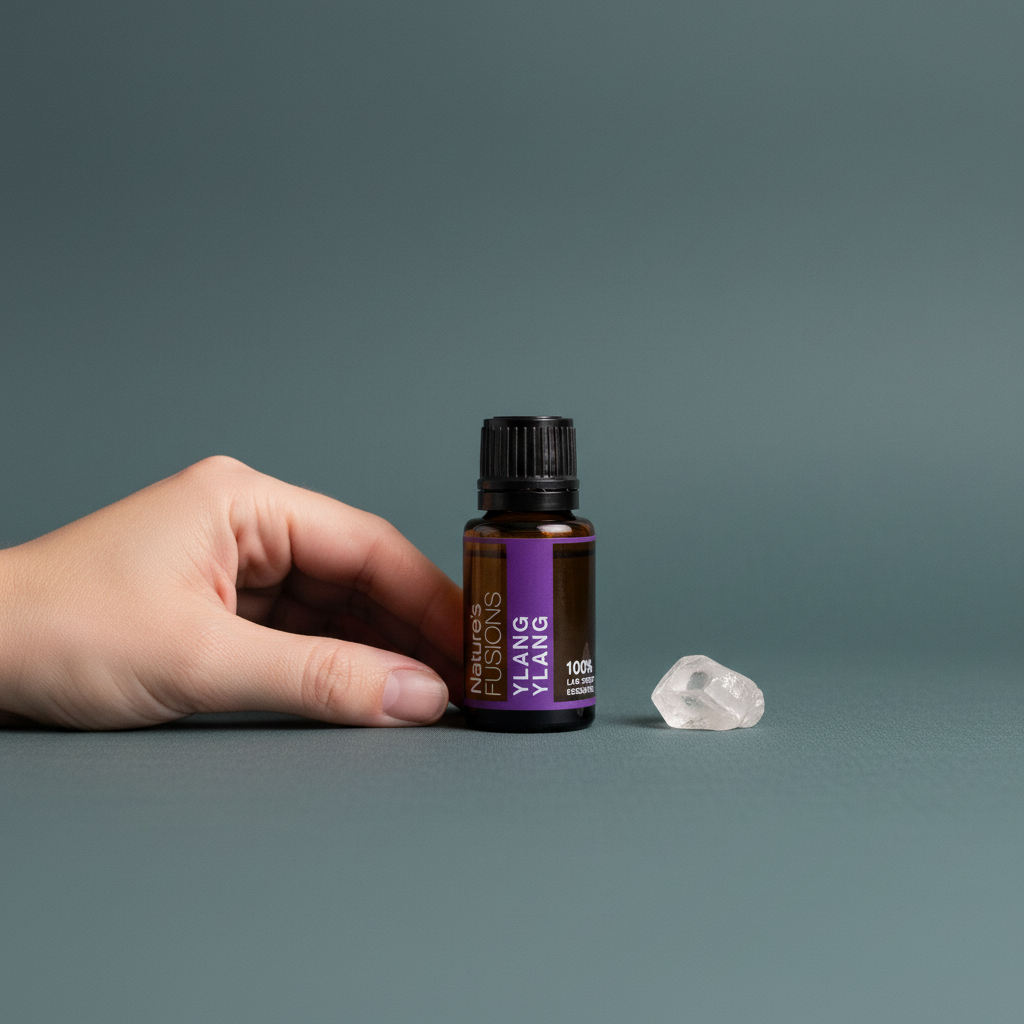 A 15ml amber glass bottle of Ylang Ylang essential oil with a black cap and purple label shown beside a human hand and a small clear crystal to demonstrate size against a muted teal background. — for Carpe Diem Dream
