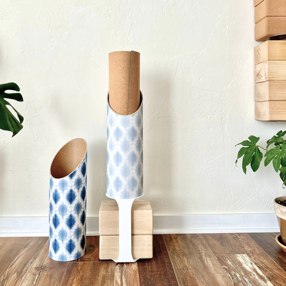 Navy and light blue patterned yoga storage tubes displayed on a wood floor with a white stand, wooden yoga blocks, plants, and a textured white wall. — for Carpe Diem Dream